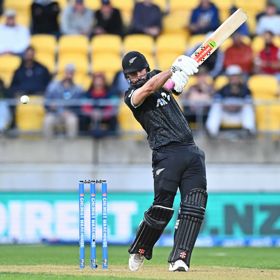 A cricket player swings a bat during a match, with stadium seating and spectators visible in the background.