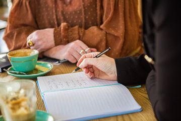 Two people are siting together, drinking coffees and writing in an open notebook.