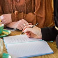 Two people are siting together, drinking coffees and writing in an open notebook.