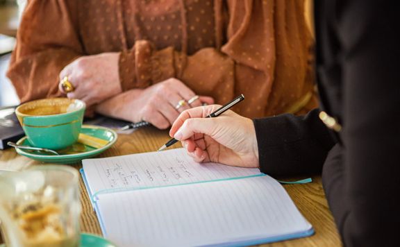 Two people are siting together, drinking coffees and writing in an open notebook.