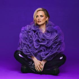Comedian, Joanne McNally, sits cross-legged wearing a purple lufa as a top and black leather pants against a purple background.
