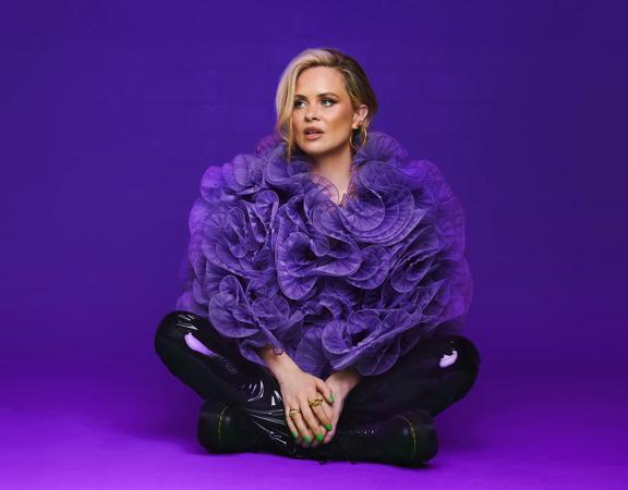 Comedian, Joanne McNally, sits cross-legged wearing a purple lufa as a top and black leather pants against a purple background.