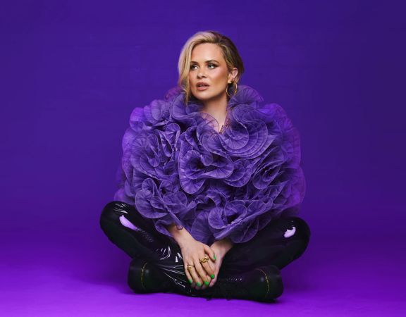 Comedian, Joanne McNally, sits cross-legged wearing a purple lufa as a top and black leather pants against a purple background.