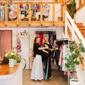 Two shoppers look through a clothing rack while a staff member stands behind the counter in a bright cozy secondhand shop.