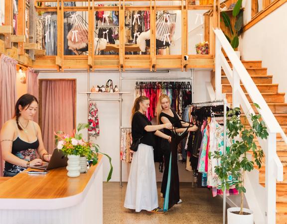 Two shoppers look through a clothing rack while a staff member stands behind the counter in a bright cozy secondhand shop.