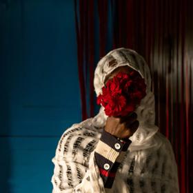 A 32-year-old Ethiopian mother poses holding a small bouquet of red flowers to hide their face in a room with dark blue walls and a sheer red curtain in the background.