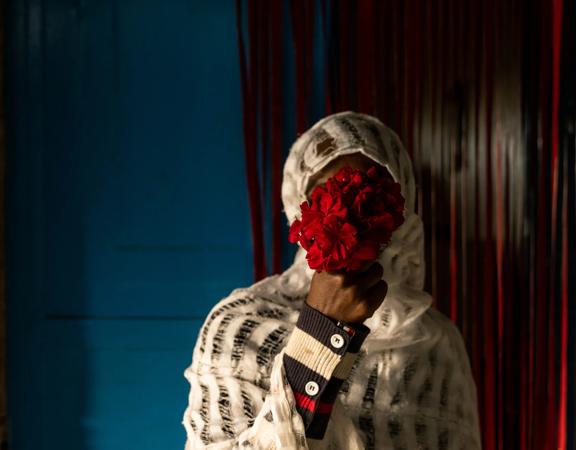 A 32-year-old Ethiopian mother poses holding a small bouquet of red flowers to hide their face in a room with dark blue walls and a sheer red curtain in the background.