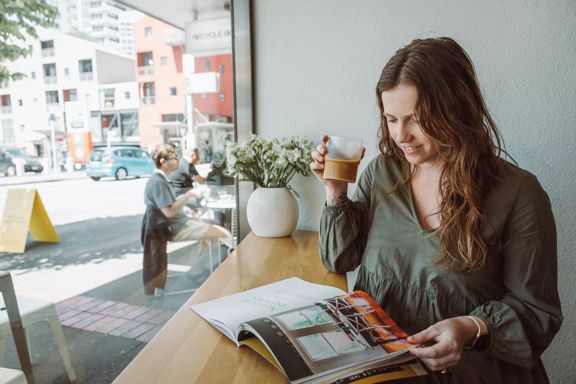 A brunette wearing a green top drinks a glass of iced coffee while reading a magazine at Customs, a cafe in Te Aro Wellington.