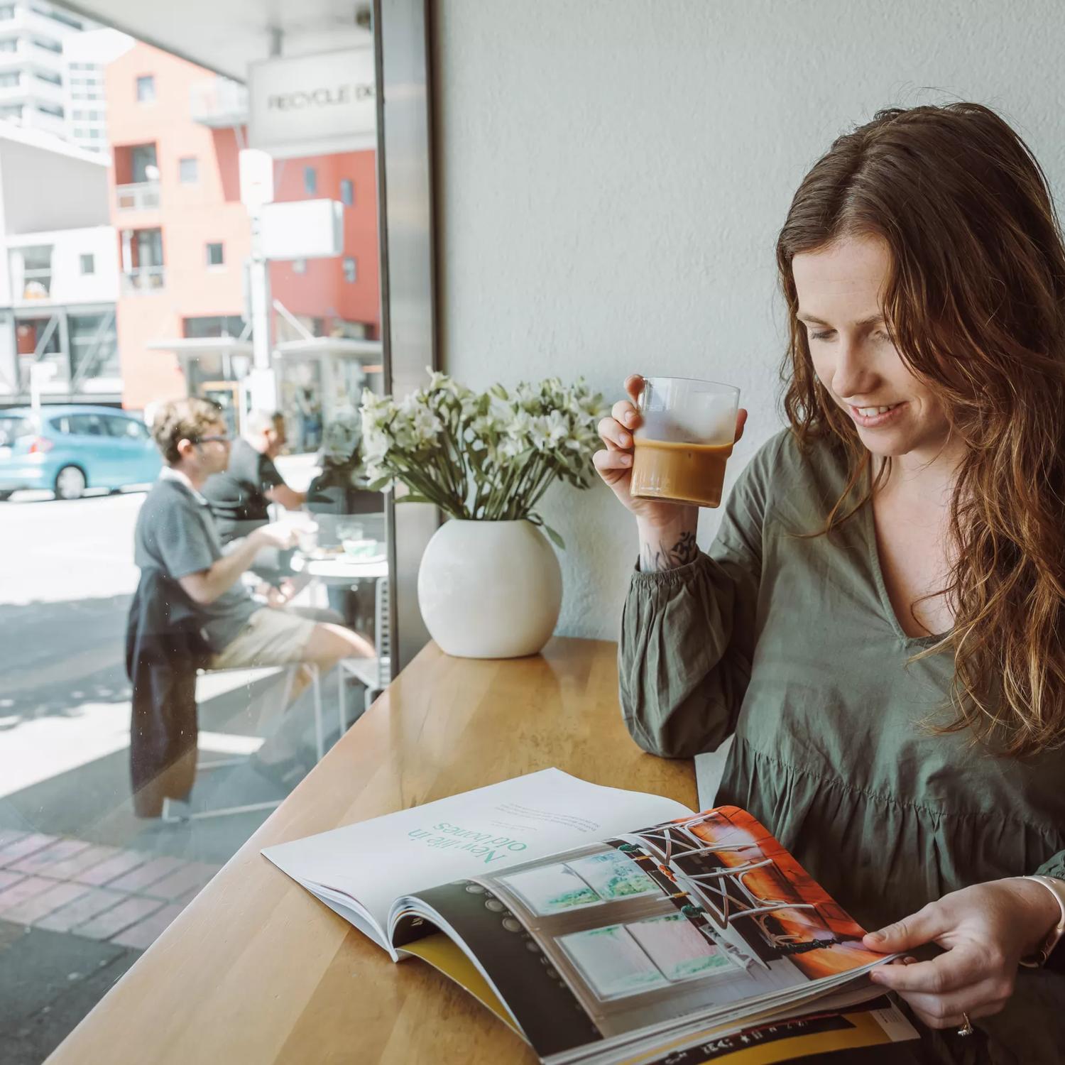 A brunette wearing a green top drinks a glass of iced coffee while reading a magazine at Customs, a cafe in Te Aro Wellington. 