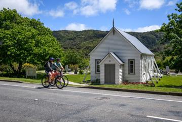 Two cyclists riding side by side on the road outside a small white weatherboard building.