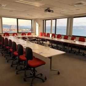 A conference room set up with tables in a U-shape, red office chairs, pens, note pads and water glasses with a view of a harbour outside the windows.
