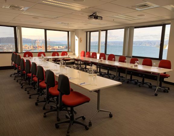 A conference room set up with tables in a U-shape, red office chairs, pens, note pads and water glasses with a view of a harbour outside the windows.