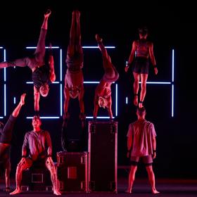 Eight aerialists pose, some do handstands, on a dark stage.