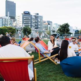 People sitting on lawn chairs and bean bags on a grassy lawn enjoying food and drinks on a lovely summers day. 