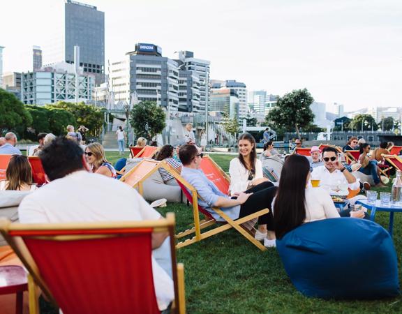 People sitting on lawn chairs and bean bags on a grassy lawn enjoying food and drinks on a lovely summers day. 