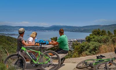 Three cyclists take a break from mountain biking. They sit at a picnic table drinking water bottles and looking at the view.