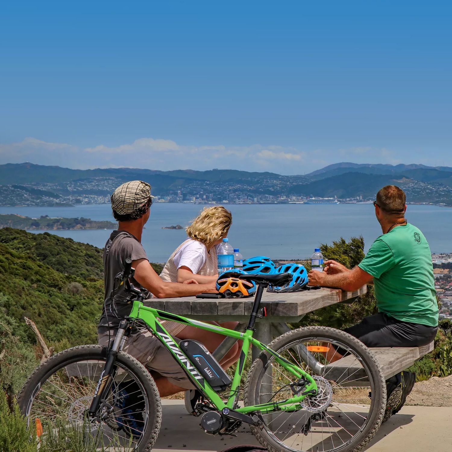 Three cyclists take a break from mountain biking. They sit at a picnic table drinking water bottles and looking at the view.