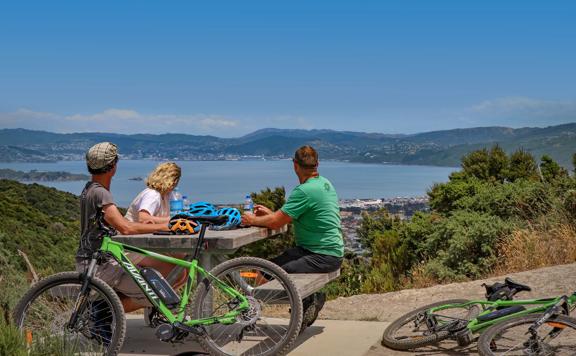 Three cyclists take a break from mountain biking. They sit at a picnic table drinking water bottles and looking at the view.