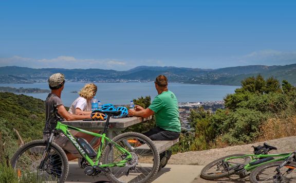 Three cyclists take a break from mountain biking. They sit at a picnic table drinking water bottles and looking at the view.