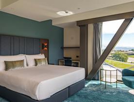 A room inside The Sebel Lower Hutt, with teal carpets and walls, a large bed and view over the Hutt with mountains in the distance.