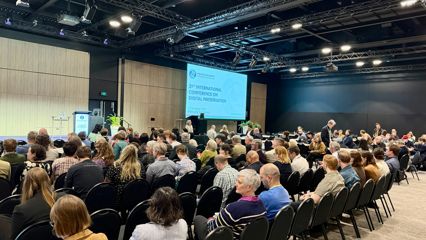 A wide shot shows a large audience seated in rows at a conference, facing a stage with a projected presentation slide at the front of the room.