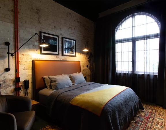 A moody and dark hotel room inside The Intrepid Hotel on Ghuznee Street. A beige brick wall is surrounded by rustic accents and a double bed.