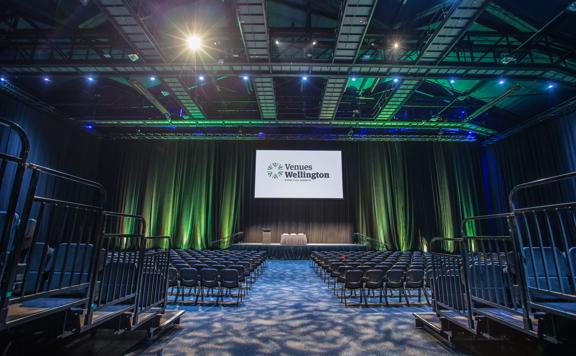 An auditorium setup at TSB Arena with a stage, green accent lighting, a projection screen, chairs and rafters. 