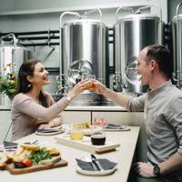 Two smiling people sit across from one another and raise their glasses to cheers. A charcuterie board, a flight of beers, fries and four small sharing plates are on the table. There are large, metal fermentation tanks behind them.