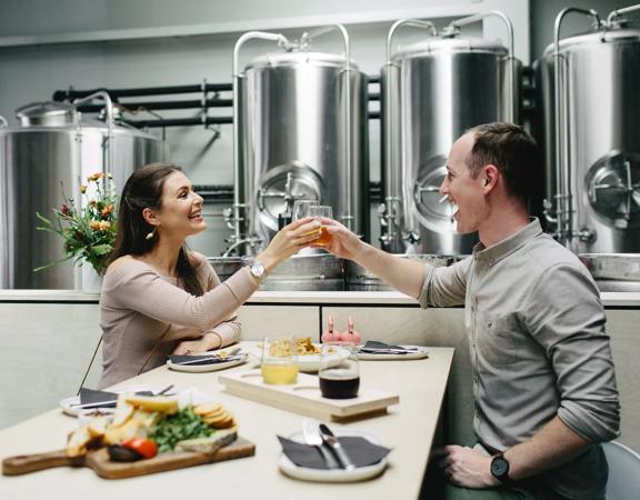 Two smiling people sit across from one another and raise their glasses to cheers. A charcuterie board, a flight of beers, fries and four small sharing plates are on the table. There are large, metal fermentation tanks behind them.