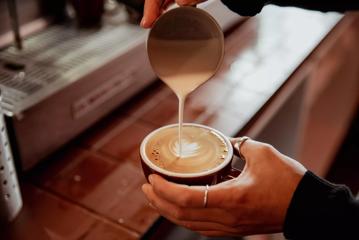Close-up on barista's hands at August Eatery in Wellington as they pour milk into a coffee cup, creating coffee art.