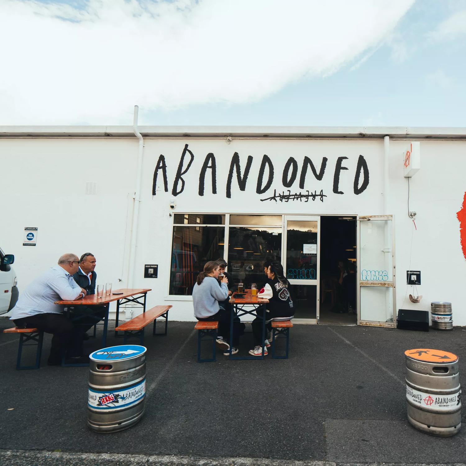 The exterior of Abandoned Taproom in Petone. It is a large white building with a large 'Abandoned' sign above the door, and the red logo on the wall. People sit in front on benches.