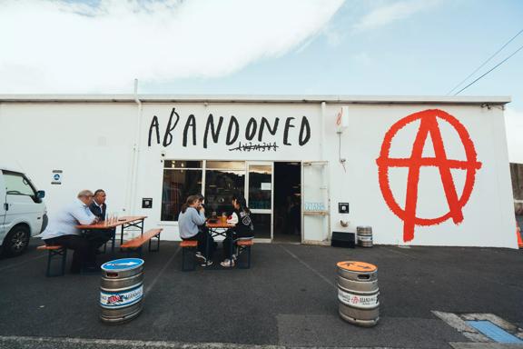 The exterior of Abandoned Taproom in Petone. It is a large white building with a large 'Abandoned' sign above the door, and the red logo on the wall. People sit in front on benches.