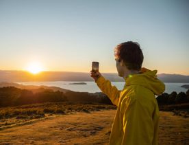 A person takes a photo of the sunrise from Mount Kaukau on Wellington’s Skyline Walkway, overlooking rolling hills and the harbour below.