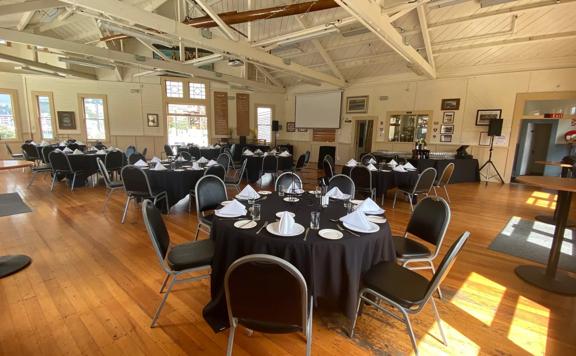 The interior of Wellington Rowing Club set up for a banquet. There are seven round tables, with black table clothes and seven chairs at each.