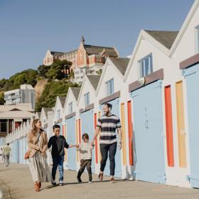 Family of 2 parents and 2 children walking along the colourful blue orange and yellow boat sheds at Oriental Bay.