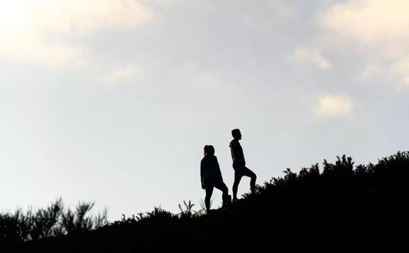 Two people are walking upwards along a grassy incline. The figures are silhouetted with a pale blue sky behind them. 