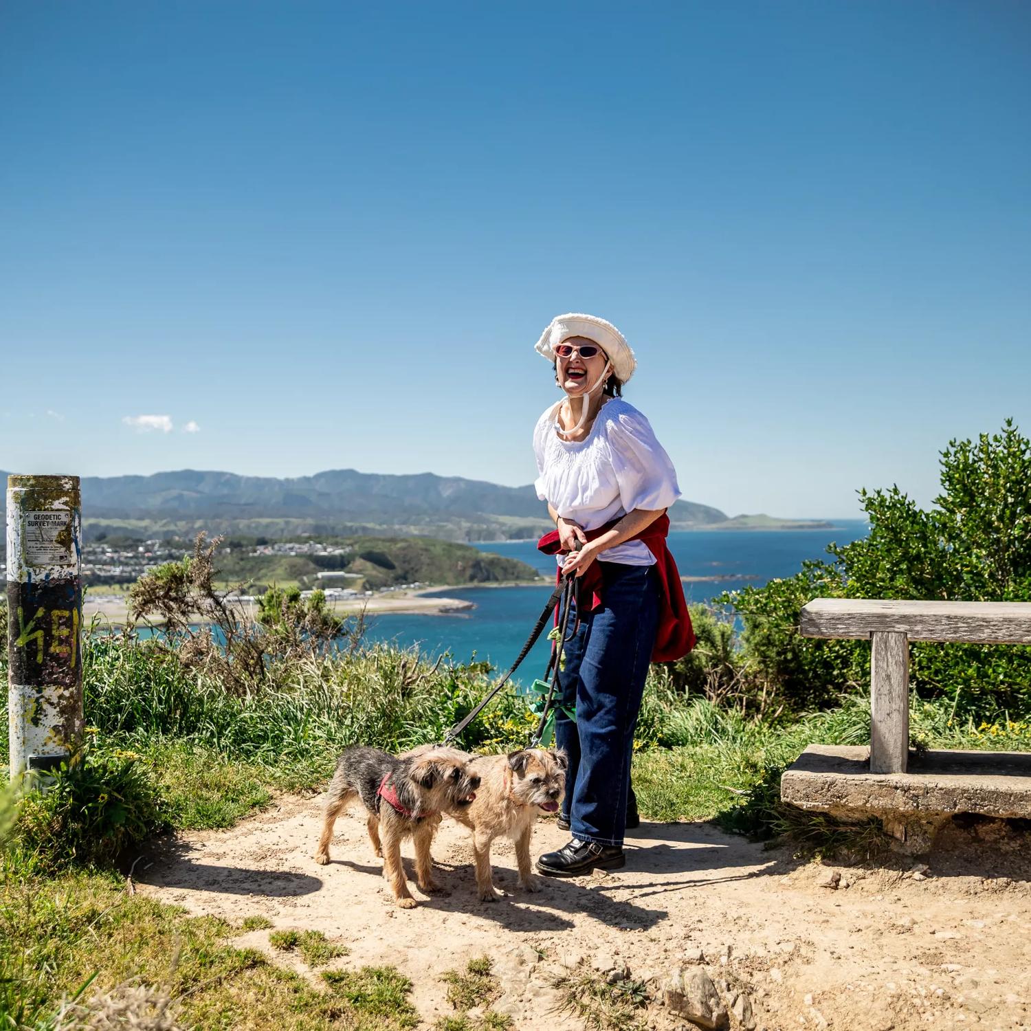 A person wearing a white top and dark blue jeans and a hat with two small dogs on a lead stands at a lookout on the Southern Walkway in the Wellington region with a scenic vista in the background.