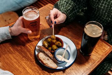 A close-up of two people drinking pints of craft beer and sharing a plate of green olives at Garage Project Taproom.