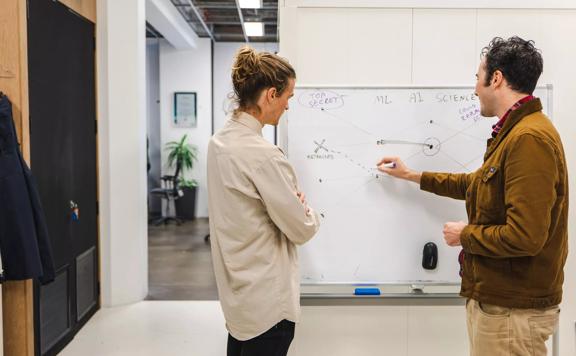 Two people drawing on a whiteboard in an office.