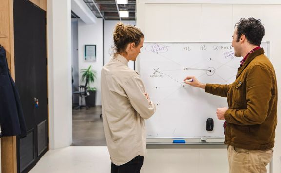 Two people drawing on a whiteboard in an office.