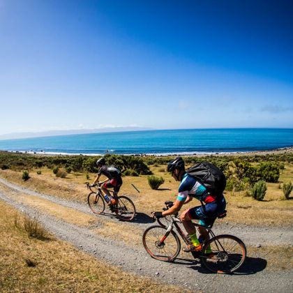 Two cyclists riding on a gravel path next to a deep blue ocean.