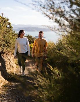 Two people walking up the Bus Barn Track in East Harbour Regional Park. Behind them, the Wellington Harbour fills the background with blue.