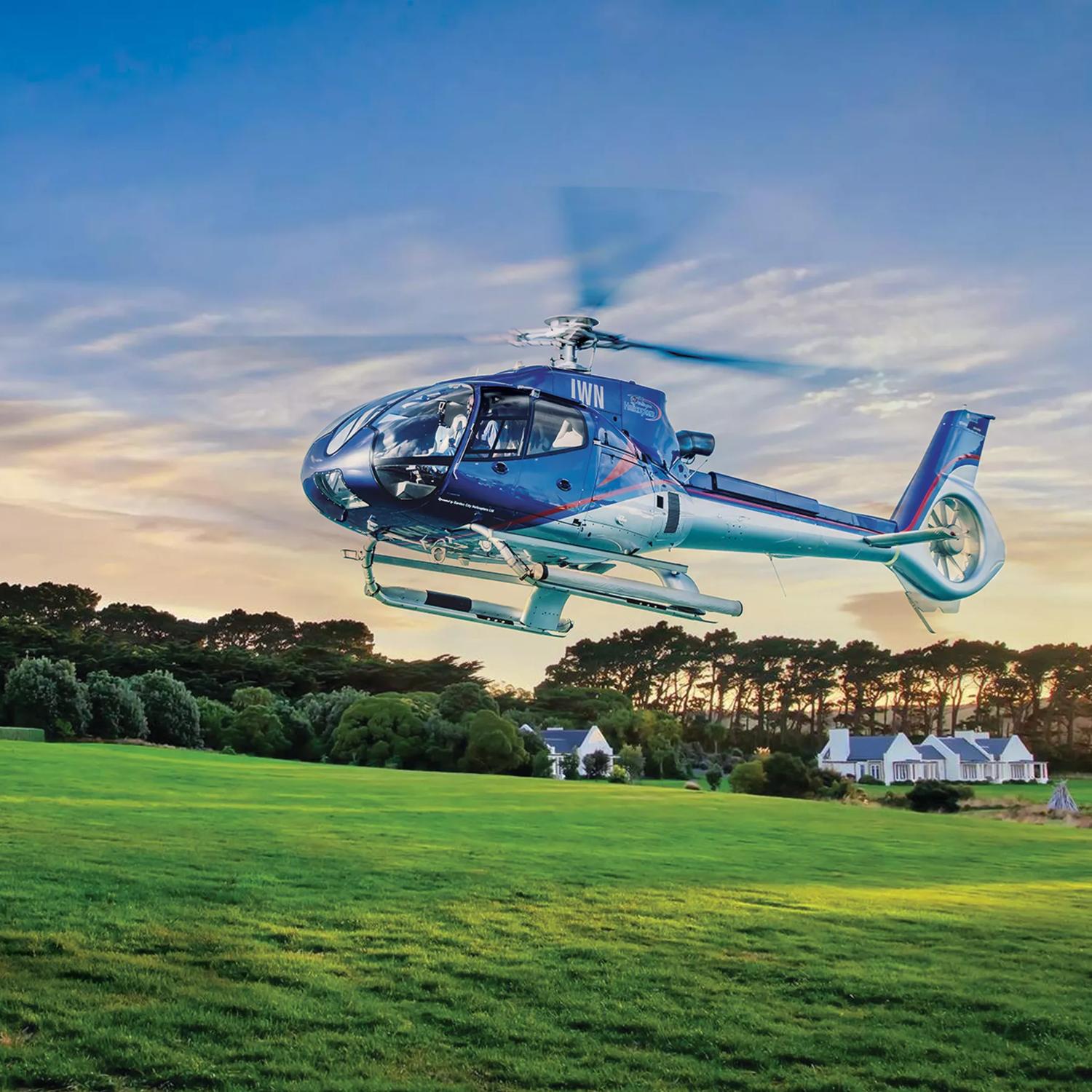 The Wellington Helicopter flying low on a field with houses in the background during sunset.