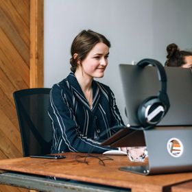 A young person sits at a desk in an industrial-style office. They're typing and looking at a monitor. A laptop sits on a stand with headphones hanging off it.