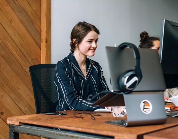 A young person sits at a desk in an industrial-style office. They're typing and looking at a monitor. A laptop sits on a stand with headphones hanging off it.