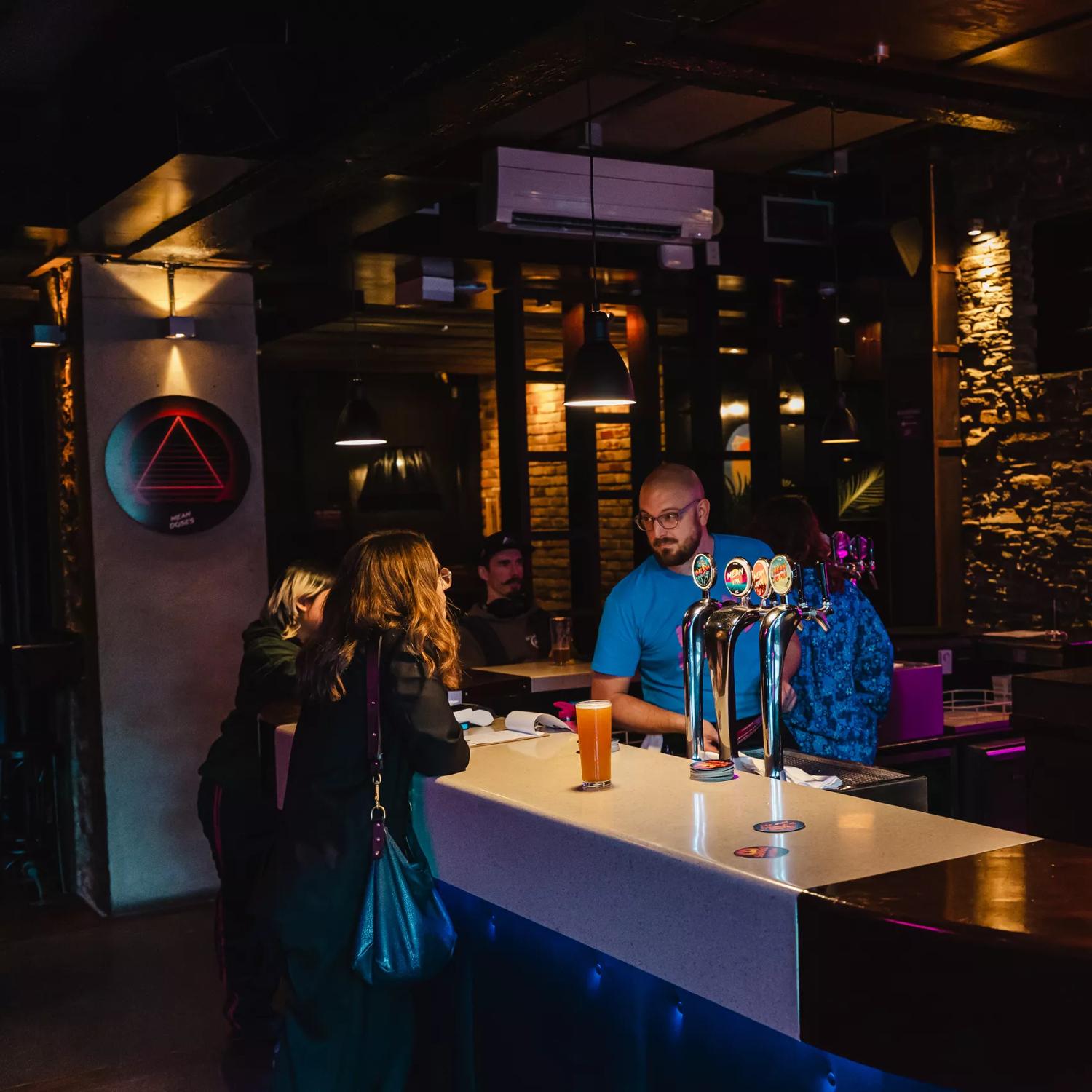 A person stands at the bar at Mean Doses Taproom, located in Te Aro, Wellington. A bartender wears a blue T shirt is working behind the counter. The space is dark with fuchsia-coloured neon lights.