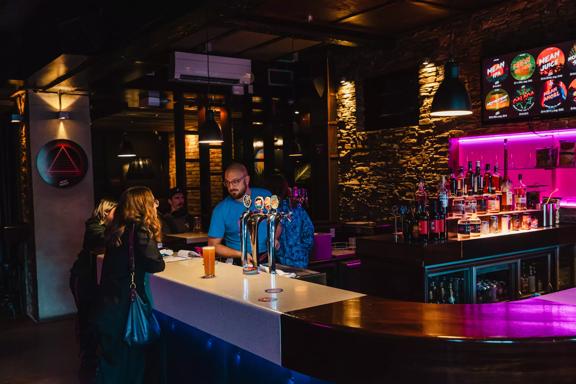A person stands at the bar at Mean Doses Taproom, located in Te Aro, Wellington. A bartender wears a blue T shirt is working behind the counter. The space is dark with fuchsia-coloured neon lights.