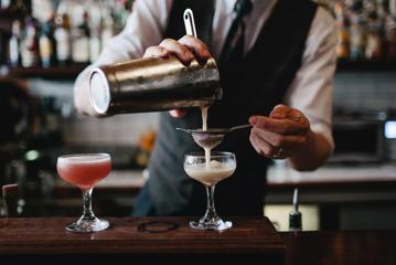 Bartender pouring a cream coloured drink into a cocktail glass, while another red drink sits beside it.