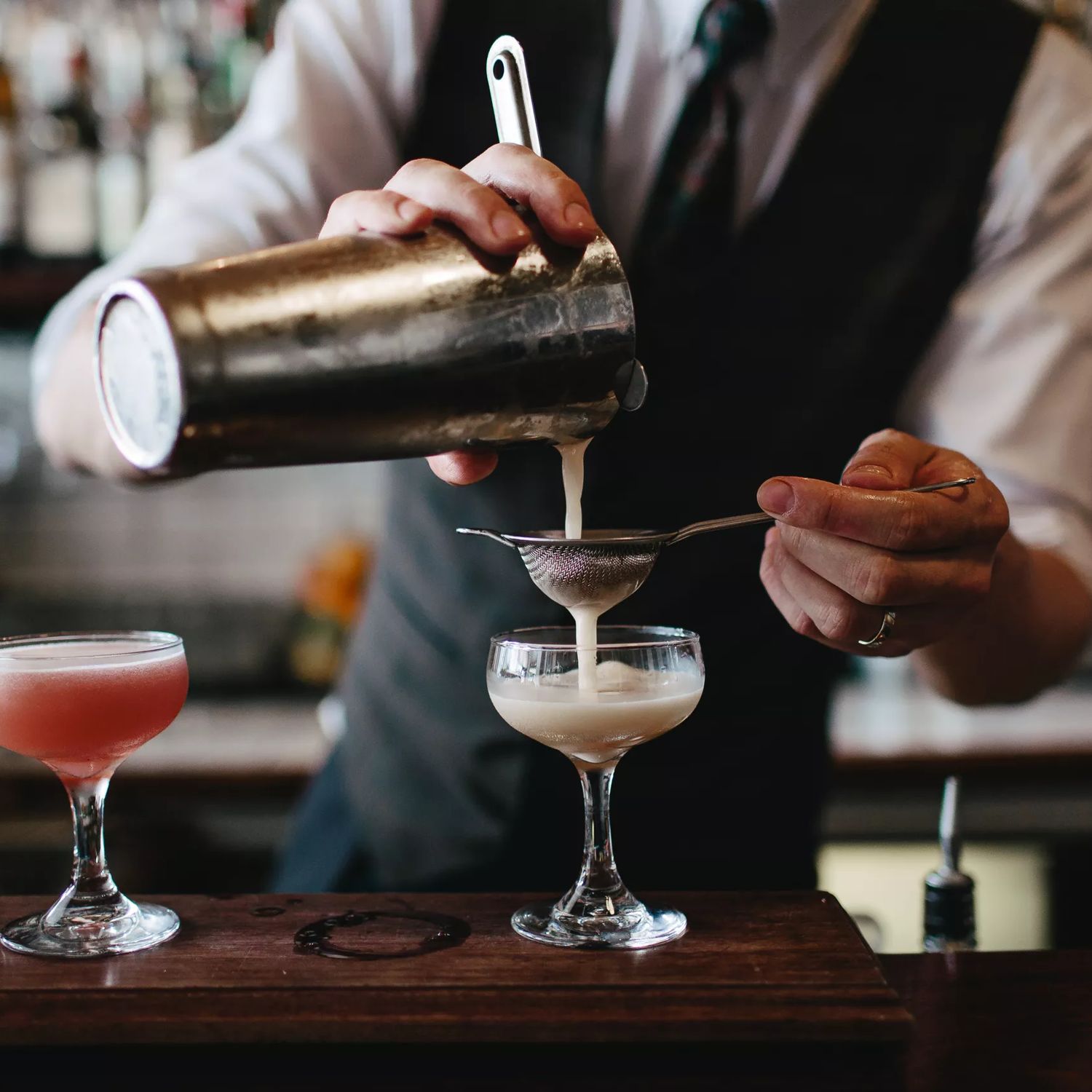 Bartender pouring a cream coloured drink into a cocktail glass, while another red drink sits beside it.