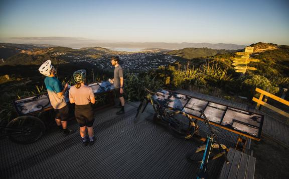 3 bikers standing on the summit of Mākara Peak Mountain Bike Park, looking over the Wellington Harbour.